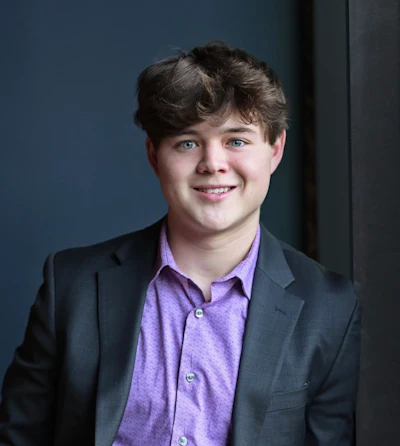 Professional headshot of young man wearing purple button-up shirt and dark blazer against blue background
