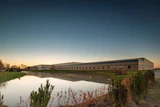 Modern industrial building complex reflected in calm water at dusk with autumn vegetation in foreground