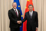 Two officials in formal business attire shake hands at a diplomatic meeting with EU and Chinese flags in the background.