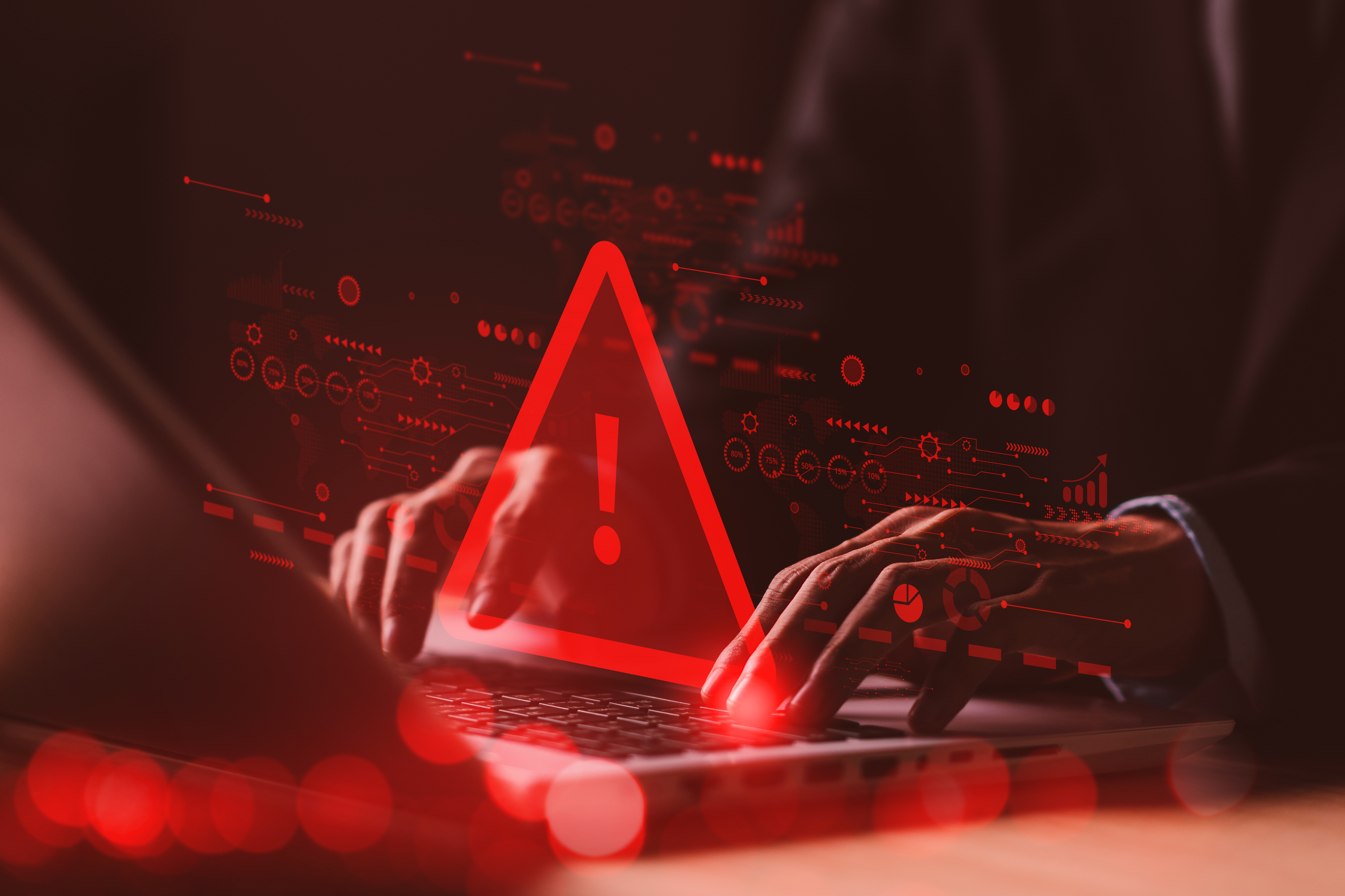 Red warning alert symbol glowing above laptop with hands, surrounded by digital data icons and circuit patterns on dark background