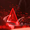 Red warning alert symbol glowing above laptop with hands, surrounded by digital data icons and circuit patterns on dark background