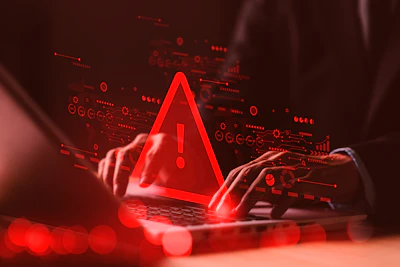 Red warning alert symbol glowing above laptop with hands, surrounded by digital data icons and circuit patterns on dark background