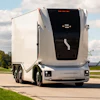 White autonomous semi-truck with black cabin parked on a paved road with green fields and cloudy sky in background