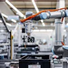 Two orange and silver robotic arms working together in a modern manufacturing facility with white structural elements and industrial equipment.