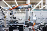 Two orange and silver robotic arms working together in a modern manufacturing facility with white structural elements and industrial equipment.