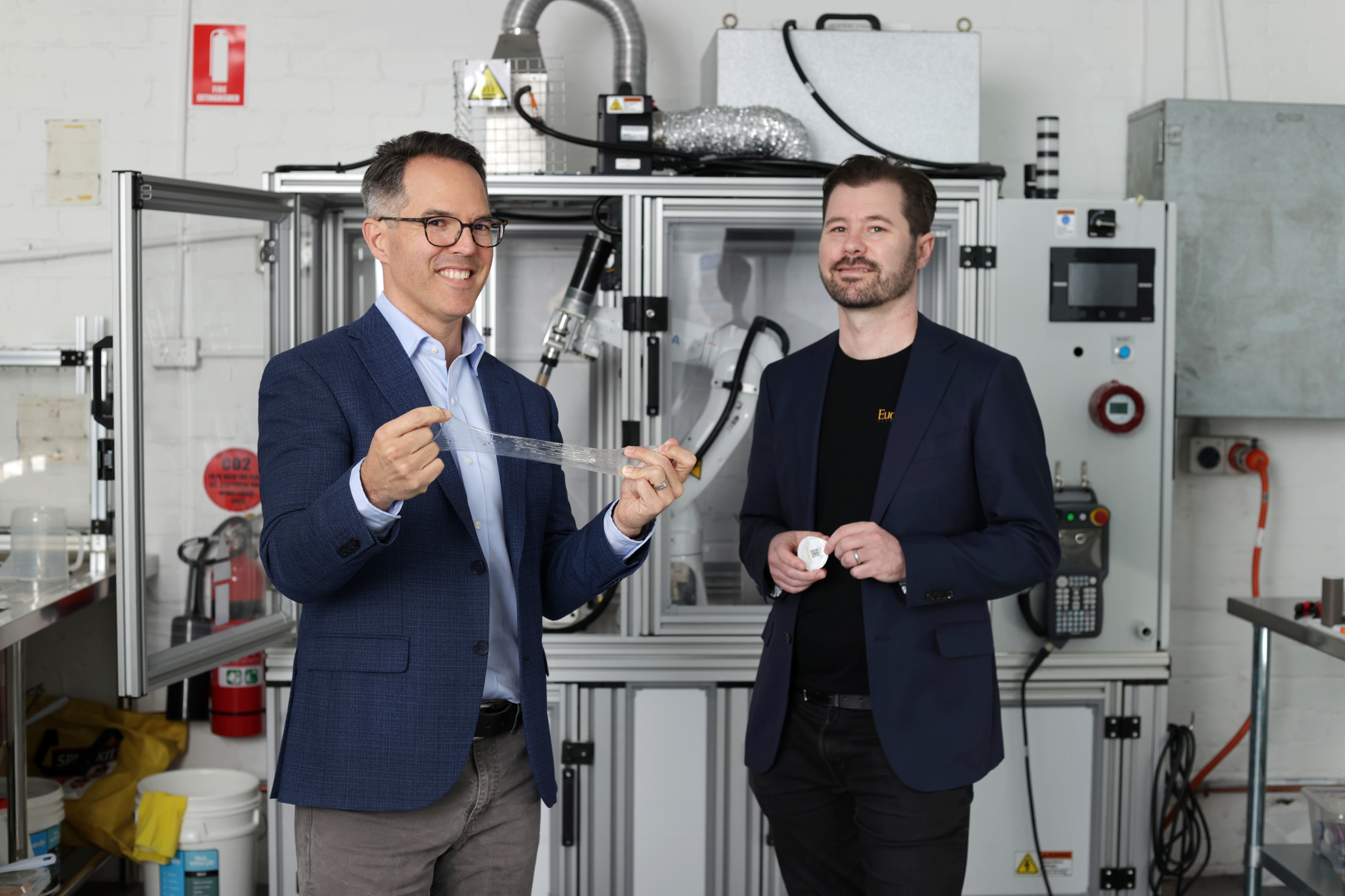 Two professionals in blazers stand in front of 3D printing equipment in a modern laboratory setting