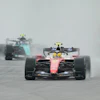 Formula 1 racing cars competing on a wet track in rainy conditions, with red and black vehicles leading the field.