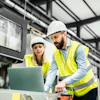 Two construction workers in hard hats and yellow safety vests review plans on a laptop inside an industrial warehouse facility.