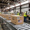 Workers in safety vests process cardboard boxes marked 'Made in USA' on a warehouse conveyor belt system.