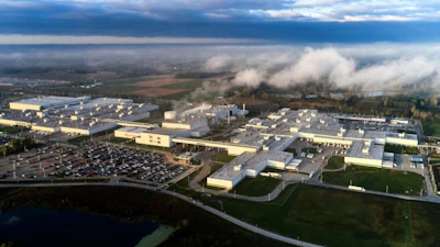 Aerial view of large industrial manufacturing complex with warehouse facilities, parking areas, and surrounding countryside landscape under cloudy sky