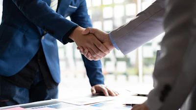 Two business professionals shaking hands over documents during a corporate meeting in a bright office setting