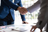 Two business professionals shaking hands over documents during a corporate meeting in a bright office setting