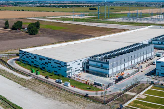 Aerial view of modern industrial warehouse complex with blue-accented buildings, loading docks, and surrounding agricultural landscape.