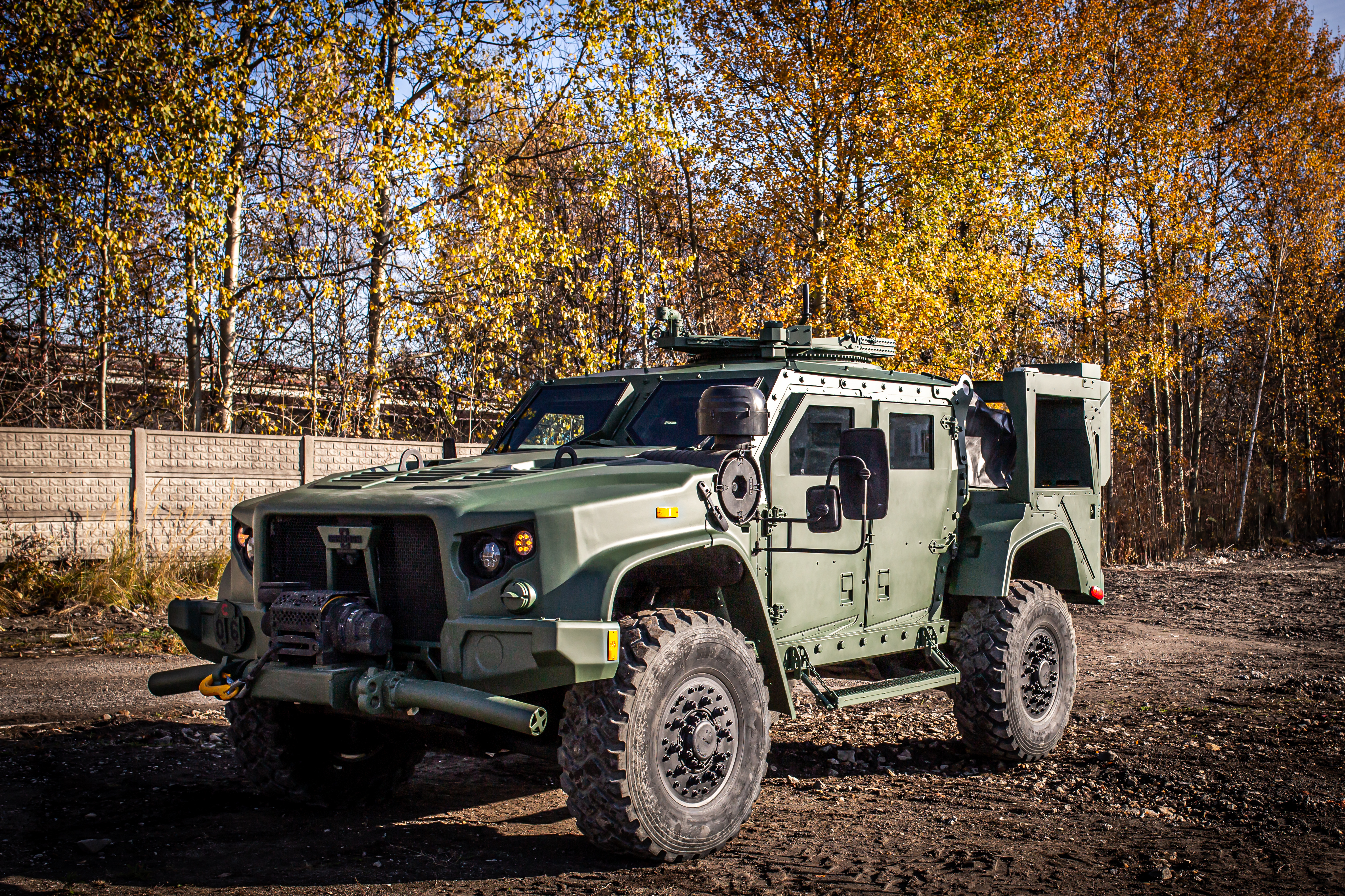 Tactical armored military vehicle with open door parked outdoors against autumn trees