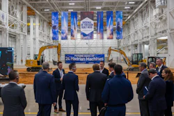 Corporate event in industrial warehouse with blue banner reading 'Future Home of 24 Hadrian Machines' and yellow excavators