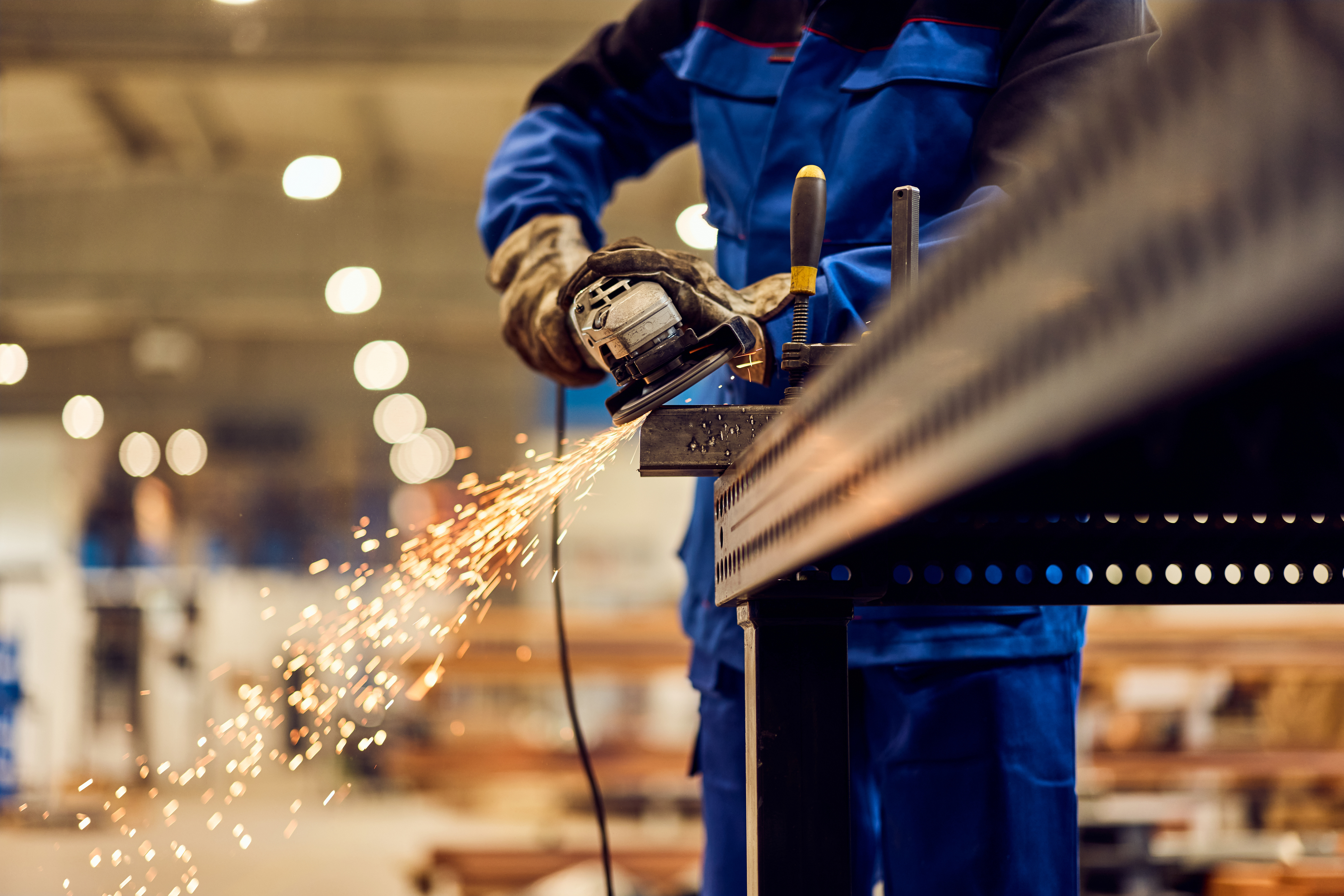 Industrial worker in blue uniform using angle grinder on metal with sparks flying in factory setting
