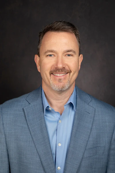 Professional headshot of a middle-aged man in blue blazer and light blue button-up shirt smiling at camera
