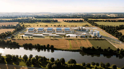 Aerial view of industrial facility with blue-roofed buildings, storage tanks, and surrounding agricultural fields near a river