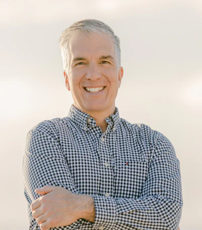 Professional headshot of a smiling middle-aged man wearing a blue and white checkered button-up shirt against a neutral background