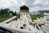White PVC pipes being unloaded from a dump truck at an industrial yard with green buildings in background