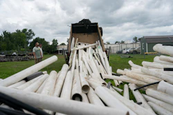 White PVC pipes being unloaded from a dump truck at an industrial yard with green buildings in background