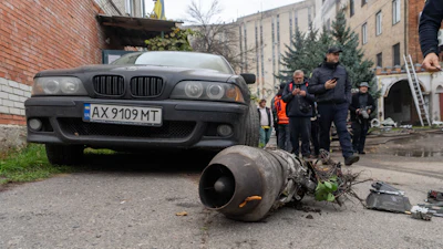 Damaged BMW sedan with detached engine component displayed on ground during investigation with officials and onlookers in urban courtyard