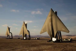 Three large metallic sail-shaped sculptures stand in a desert landscape under a blue sky with clouds