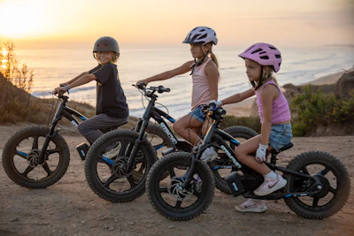 Three children wearing helmets ride bikes on a sandy beach at sunset with ocean waves in the background.