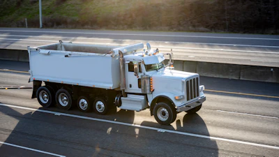 White Peterbilt dump truck traveling on a multi-lane highway with grassy hillside in background