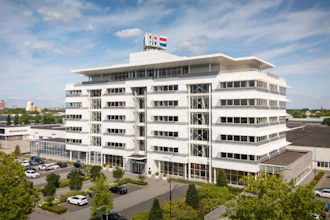 Modern white multi-story commercial building with glass windows and flags on rooftop, surrounded by landscaping and parked vehicles