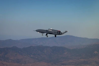 Dark aircraft in flight over mountainous terrain with clear blue sky